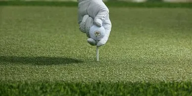 A close-up of a hand wearing a golf glove placing a golf tee into artificial golf turf.