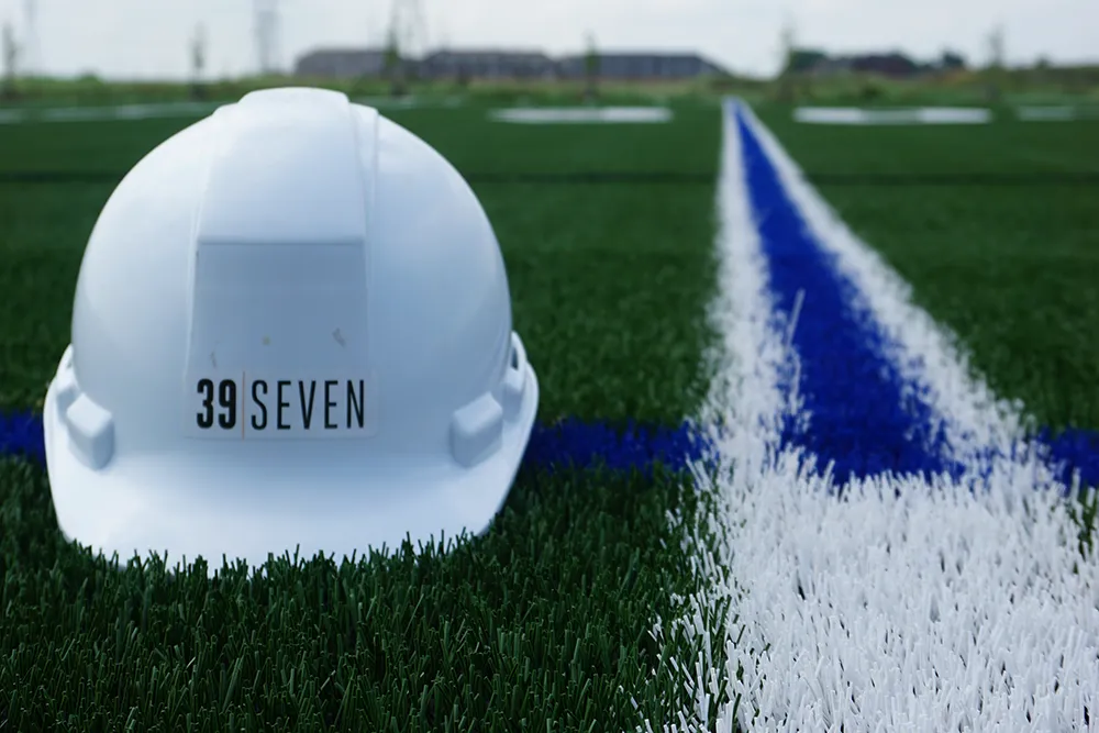 A hard hat with the 39 Seven logo lays beside a field line painted on artificial turf.